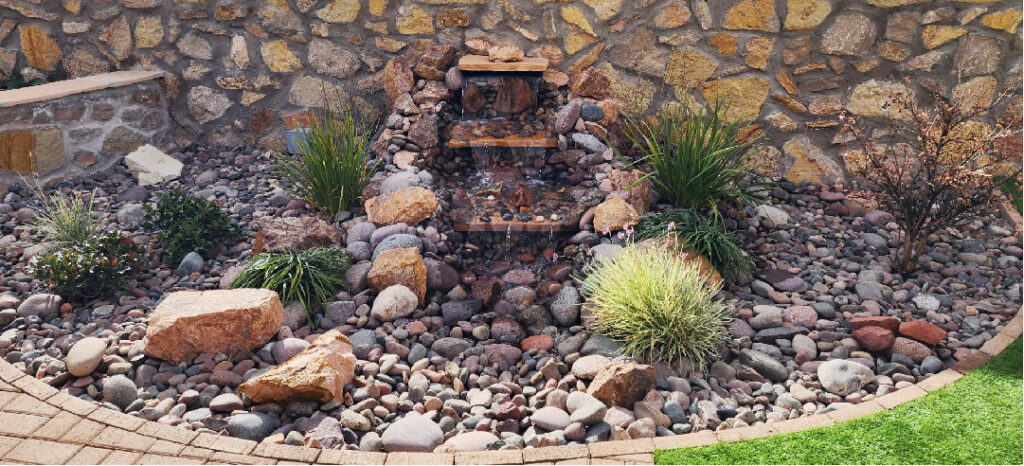 A cascading stone fountain against a stone wall is displayed working in a backyard.